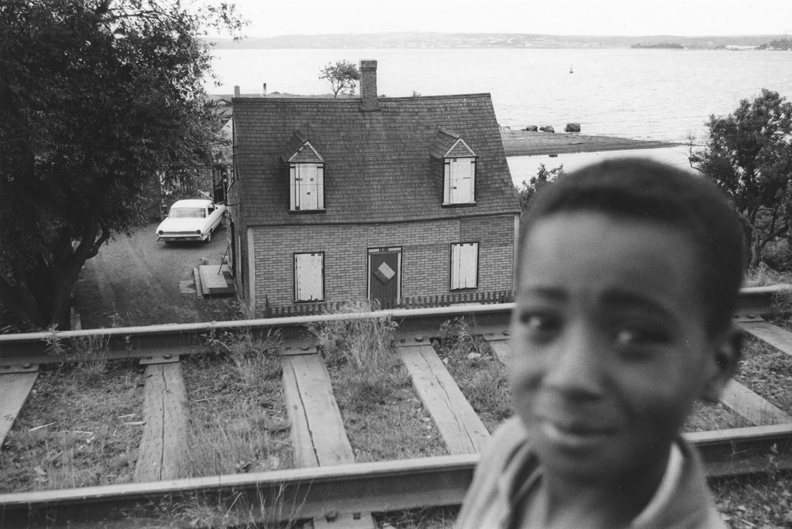 africville : Young boy, with Ralph Jones house, Africville, boarded up prior to demolition, in the background