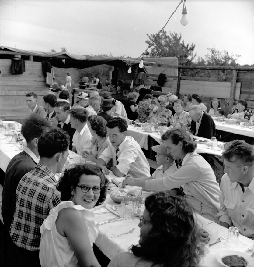 Participants eating supper at outdoor picnic tables sheltered by old ships' sails at the Catholic Church Picnic; Doucetteville; August 27, 1950  