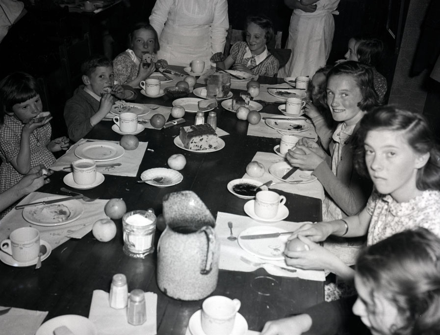 Several shots of a large group of people, including British evacuee children, eating lunch